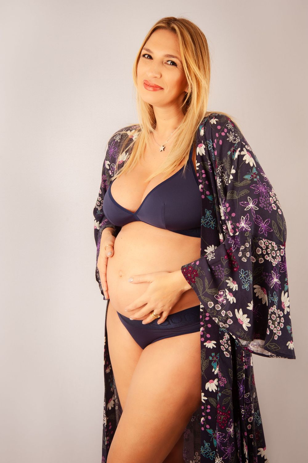 Smiling pregnant blonde woman in navy bikini and floral kimono posing for maternity photo.