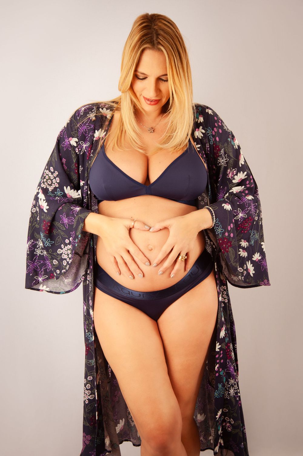 Pregnant woman in navy bikini and floral robe making heart shape with hands on belly.