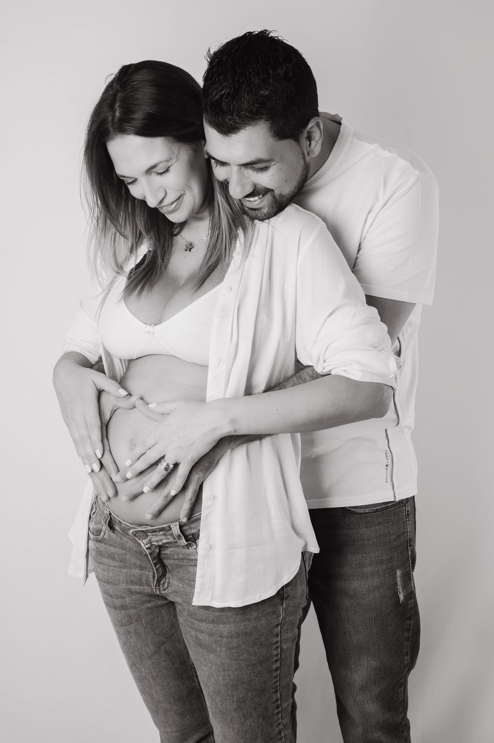 Black and white maternity shoot of smiling couple cradling pregnant belly together in white tops and jeans.