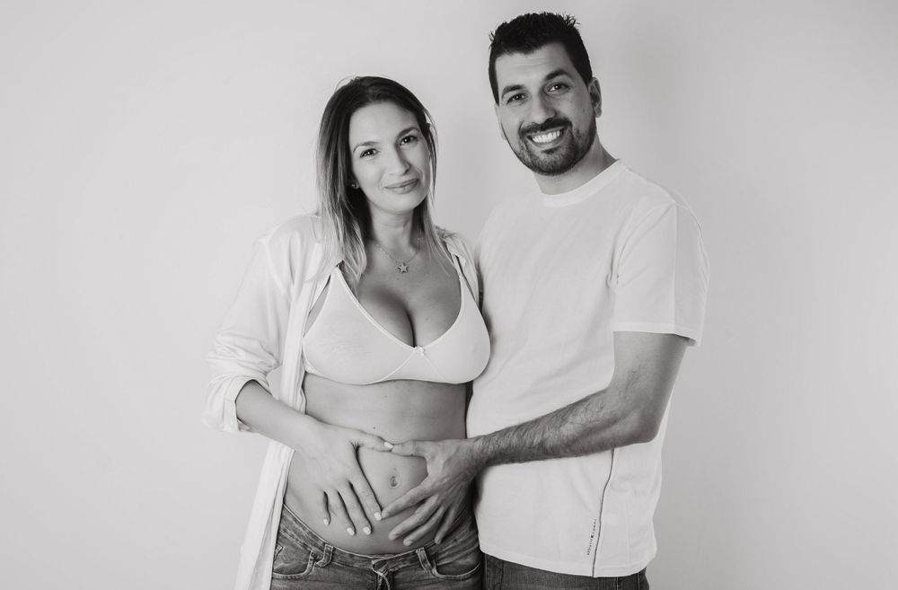 Black and white maternity photo of smiling couple posing together, hands on pregnant belly in studio setting.