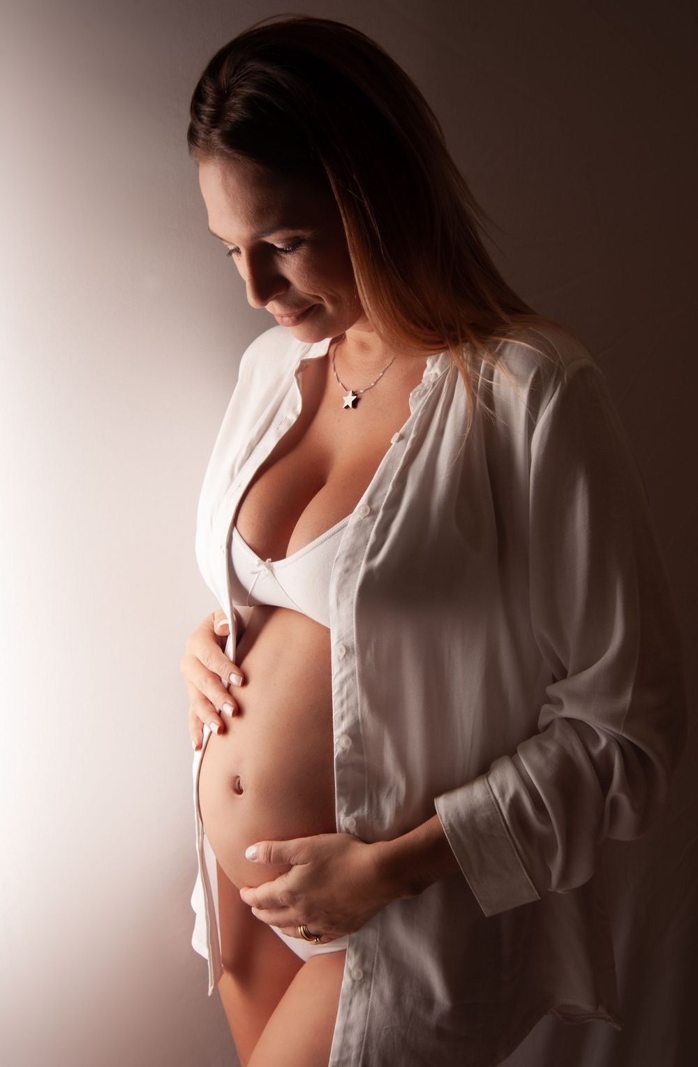 Pregnant woman in white open shirt and lingerie cradling baby bump in dramatic studio lighting portrait.