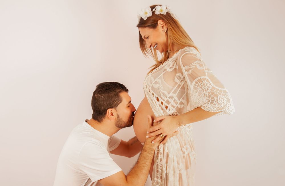 Man kneeling to kiss pregnant woman's belly during maternity photoshoot, woman wearing white lace dress with floral headband.