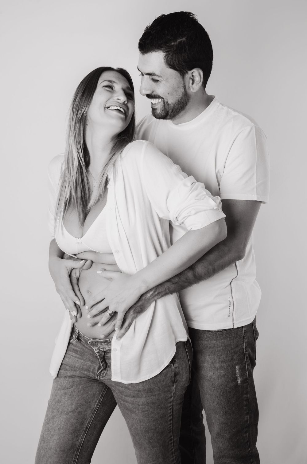 Black and white photo of happy couple embracing during maternity photoshoot in casual clothes.