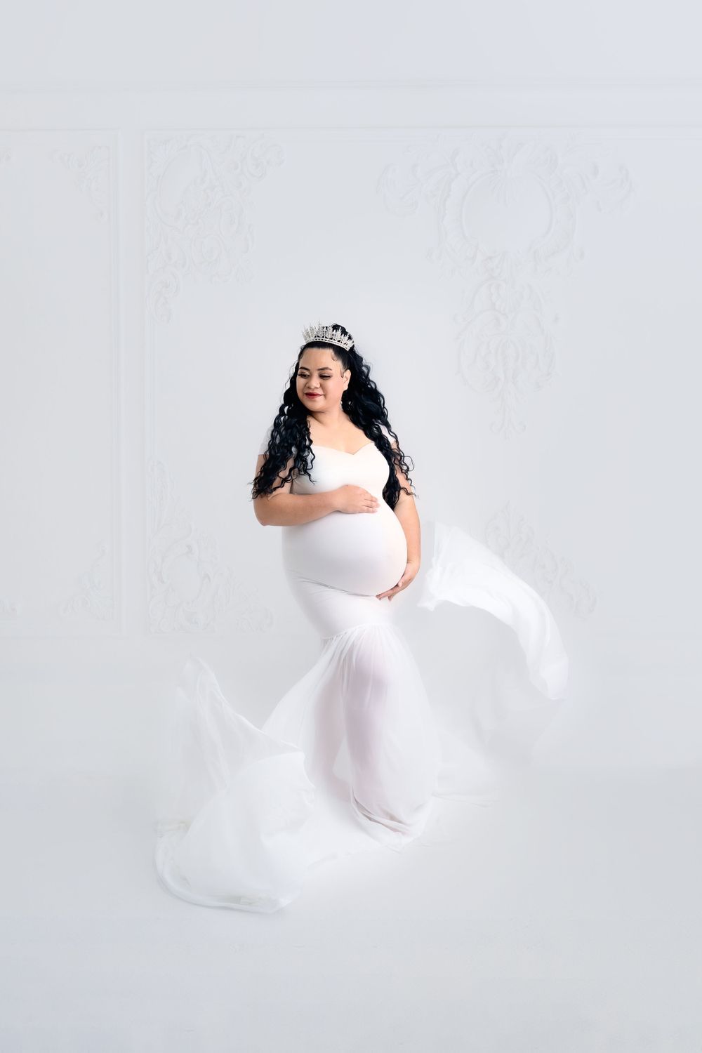 Dreamy maternity session with billowing white dress captured in motion against white backdrop.