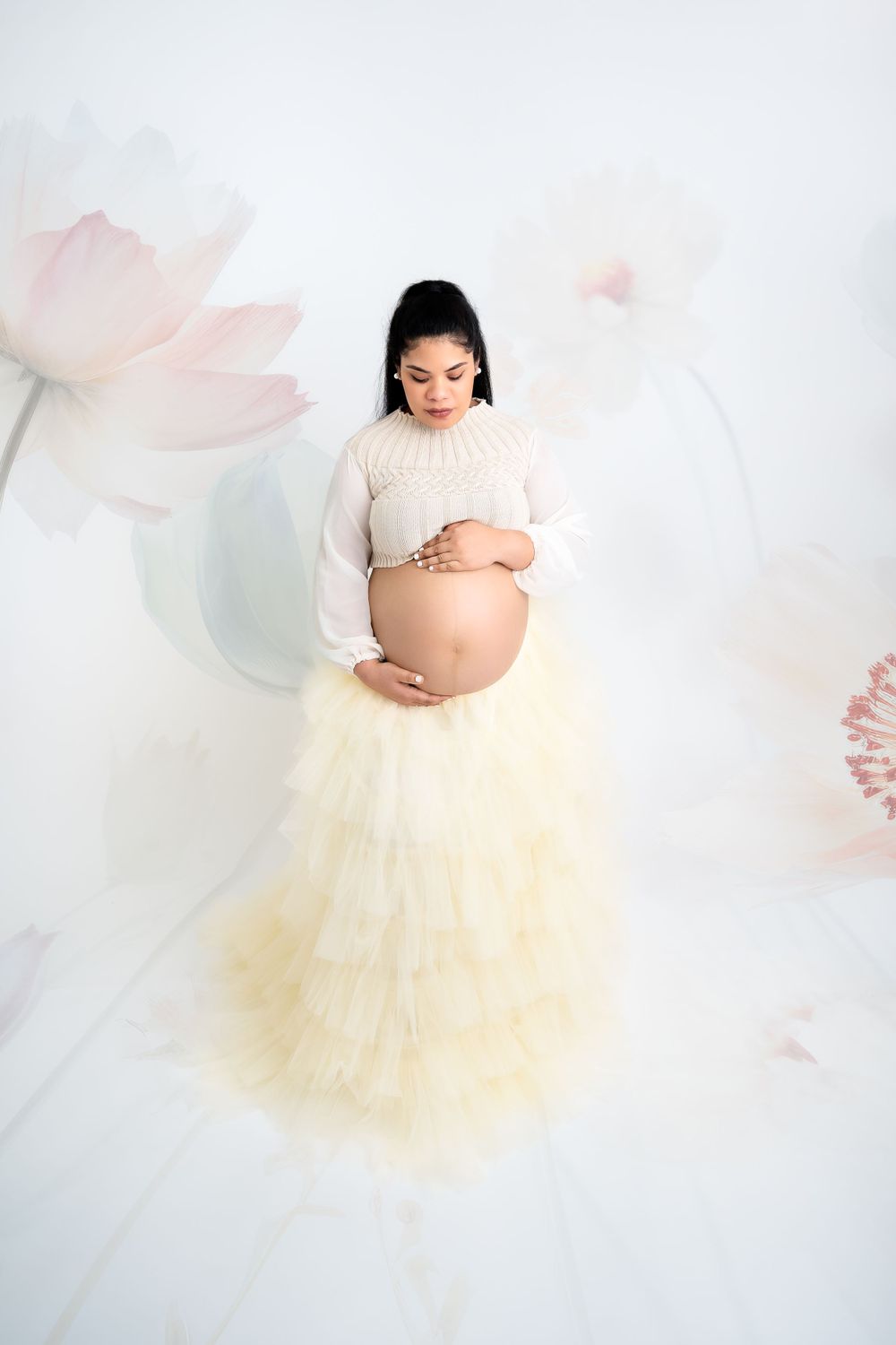 Artistic maternity portrait with cream tulle skirt against soft floral backdrop.
