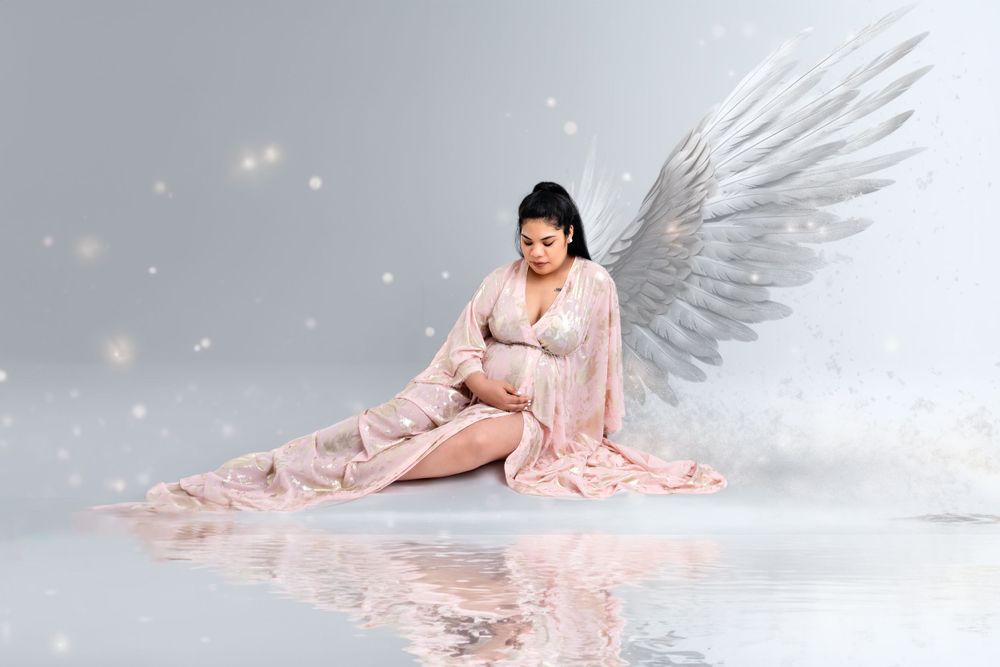 Ethereal maternity portrait with angel wings and pink dress in soft lighting.