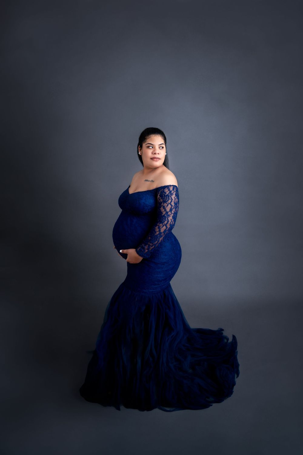 Elegant maternity photoshoot featuring a flowing navy blue dress against a dark grey backdrop.