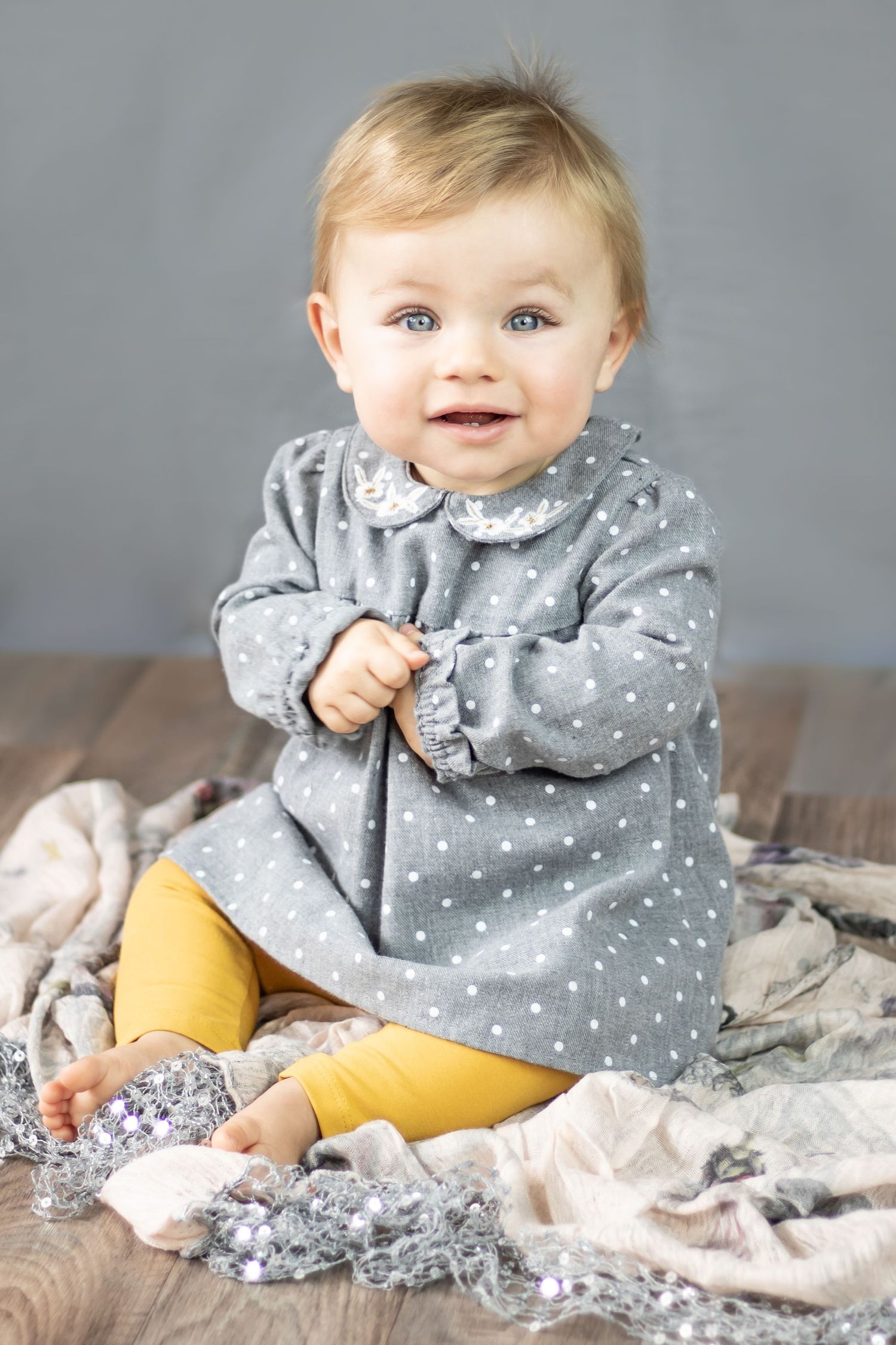 Baby sitting for a photo shoot. wearing grey spotty dress and yellow leggings. she is smiling and holding her hands