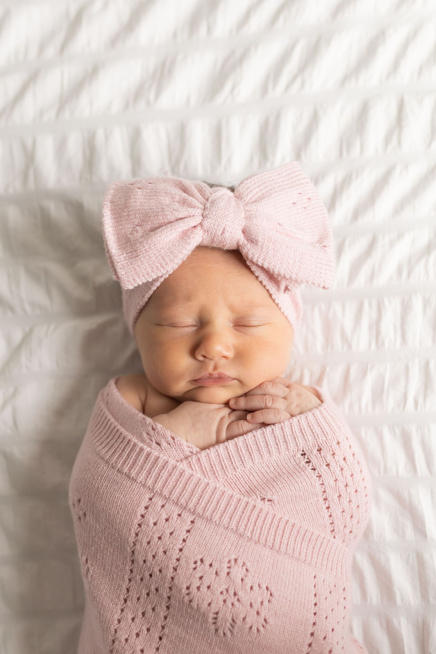 Sleeping newborn wearing a pink bow headband and knit blanket on white bedding.