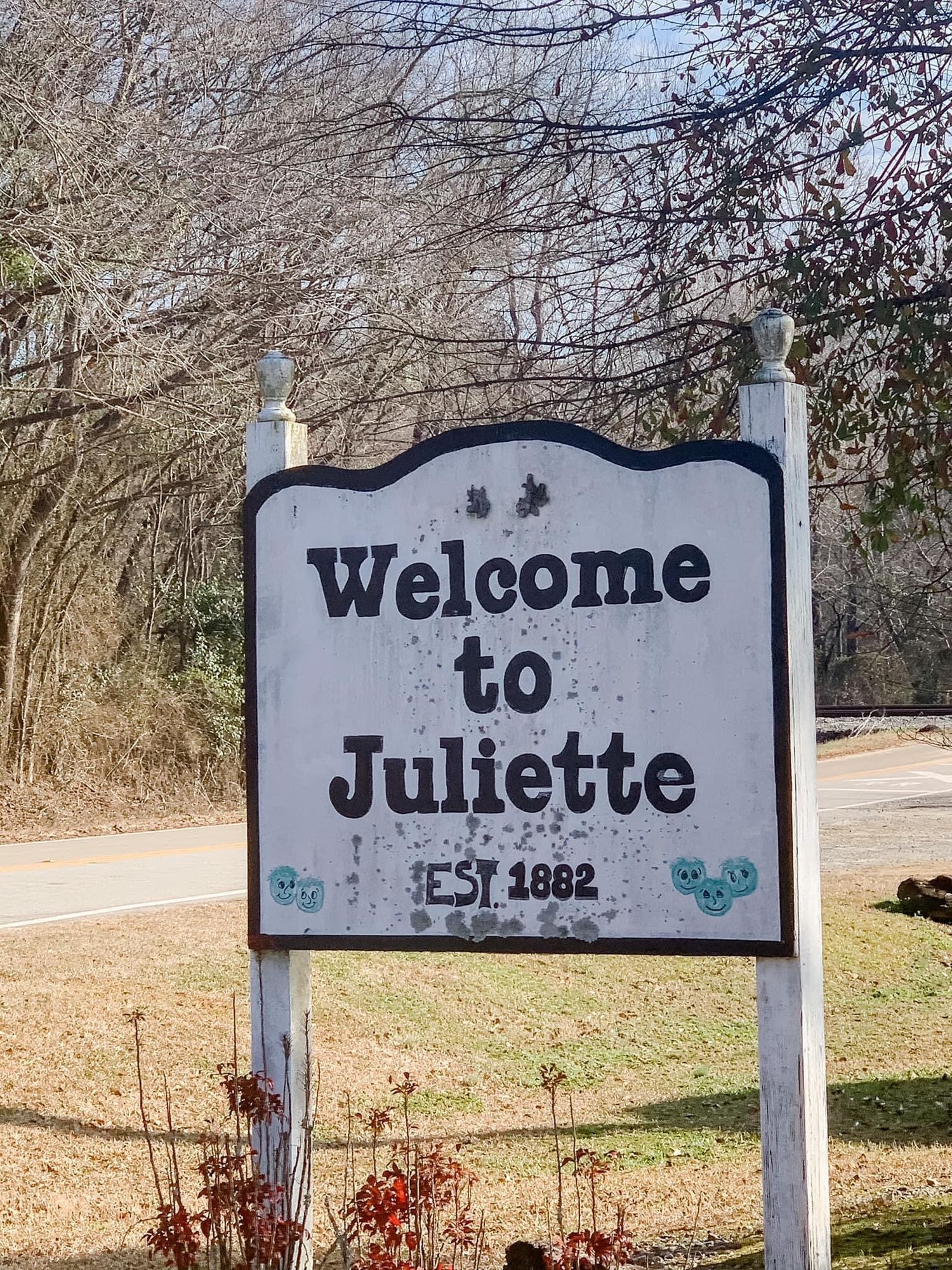 Weathered welcome sign for Juliette with decorative floral accents against winter trees.