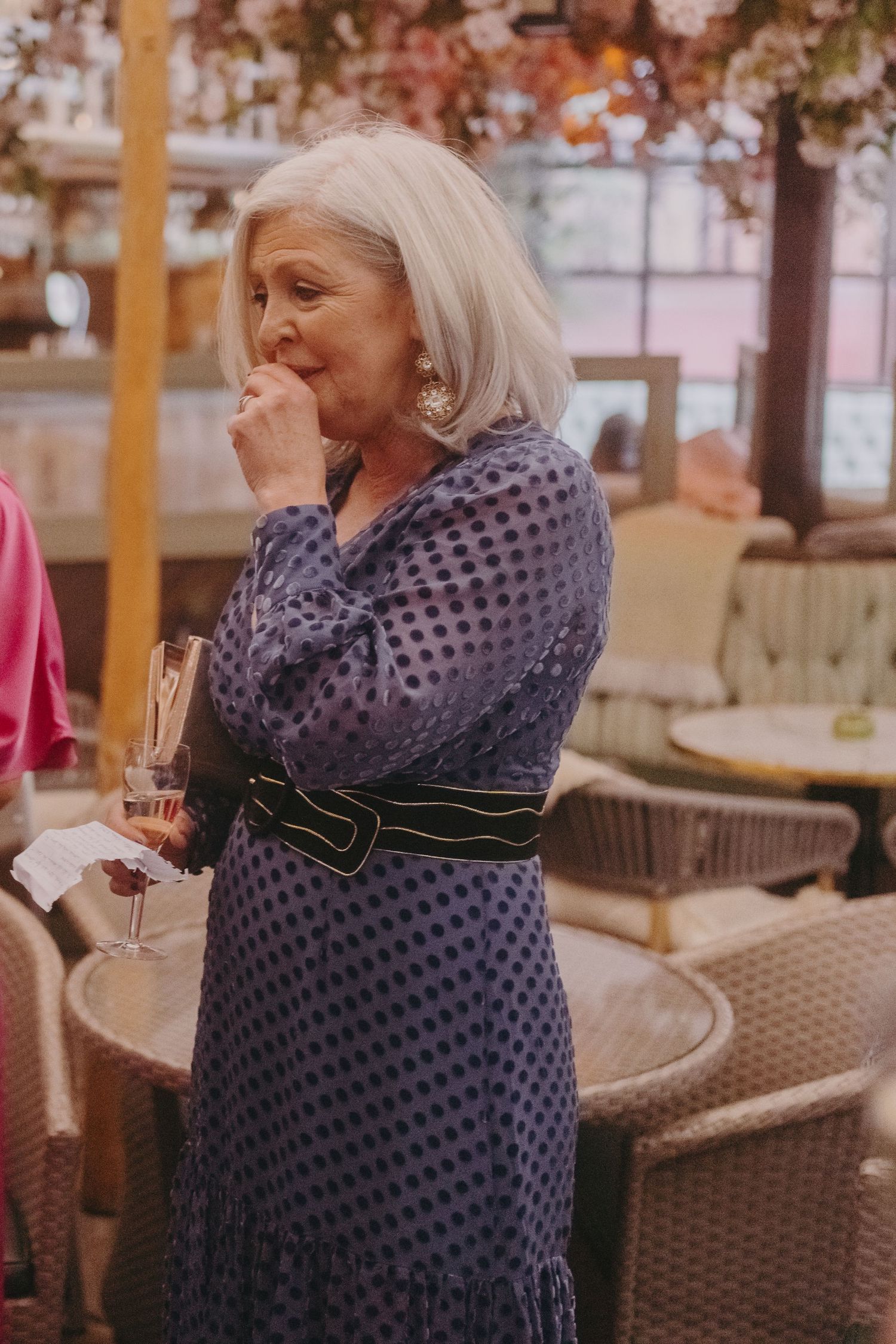 An elegant woman in a navy polka dot dress with silver hair holds a champagne glass at an outdoor event.