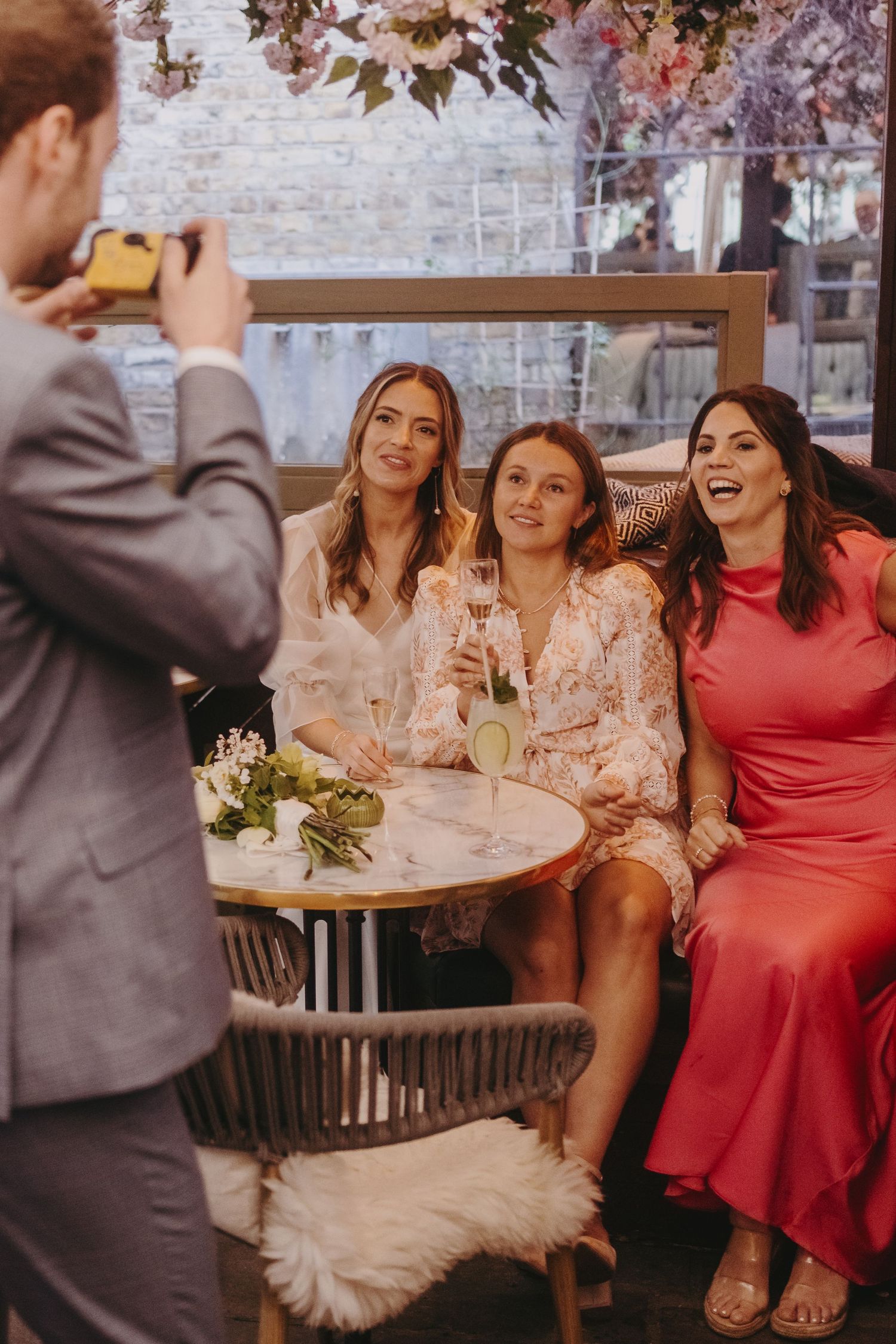 Three friends enjoying drinks and conversation at an outdoor restaurant table while being photographed.