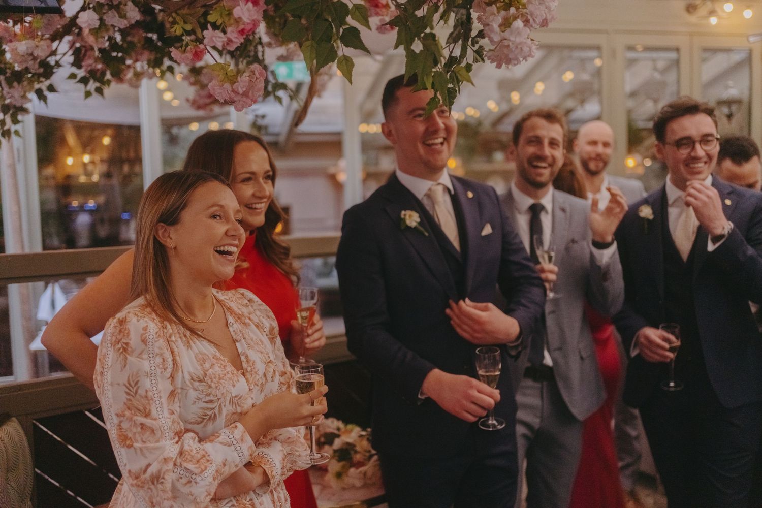 Wedding guests sharing laughter and drinks during a joyful celebration under floral decorations.