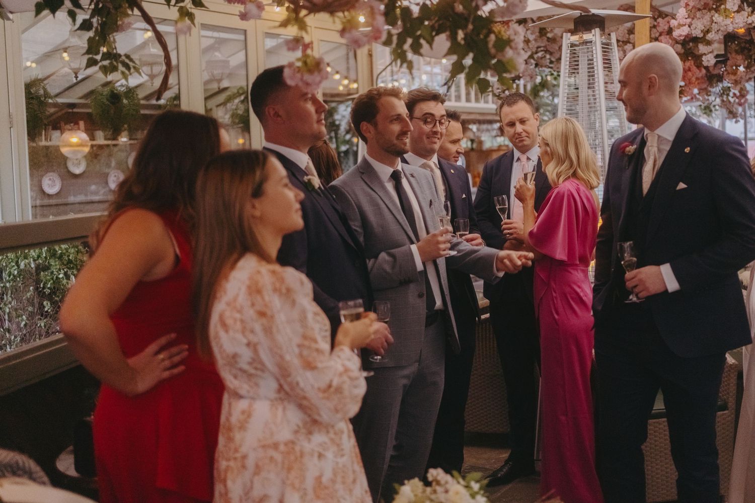Wedding party gathered indoors enjoying conversation and drinks under elegant floral arrangements.