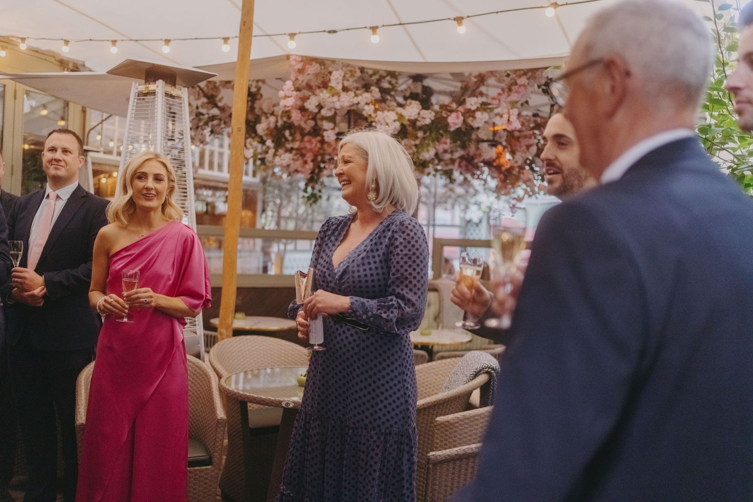 Guests in formal attire socializing at an outdoor event decorated with pink floral arrangements.