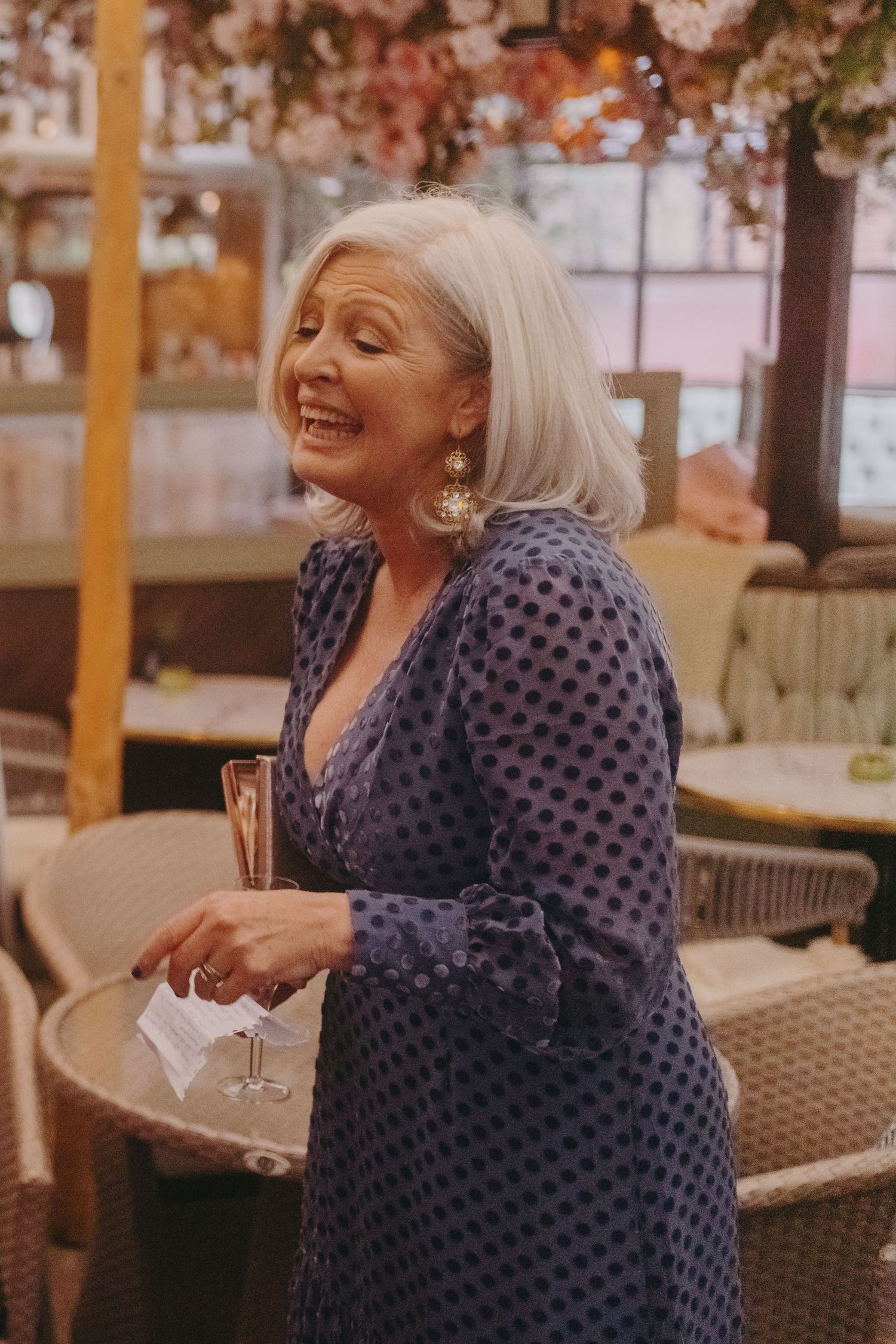 Woman wearing navy polka dot dress and statement earrings laughs while holding champagne at outdoor gathering.
