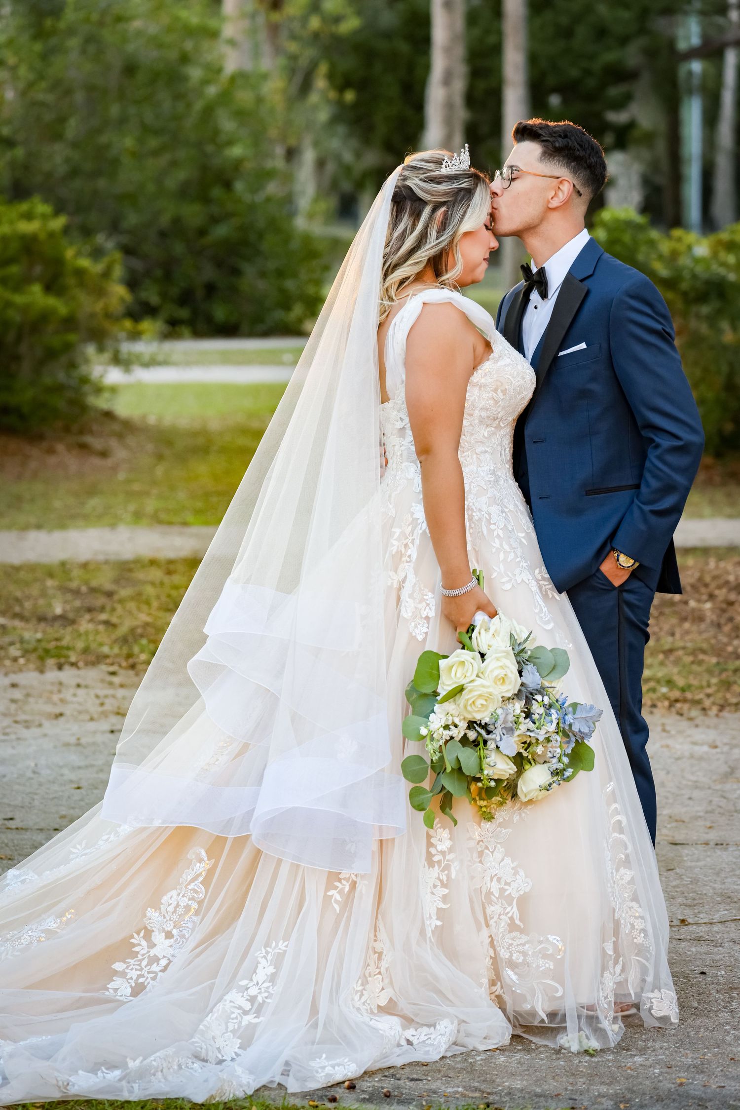 Newlywed couple sharing a kiss in a garden setting with bride holding a white and blue bouquet.