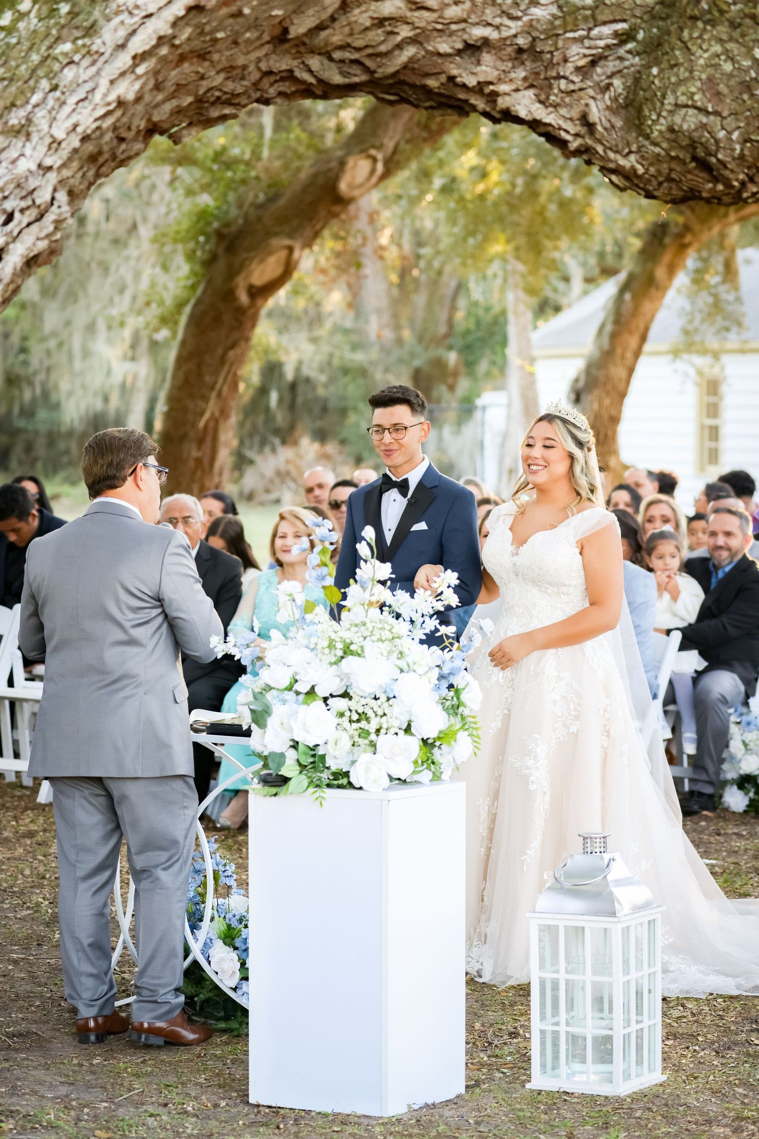 Outdoor wedding ceremony under large oak trees with white floral decorations and lanterns.