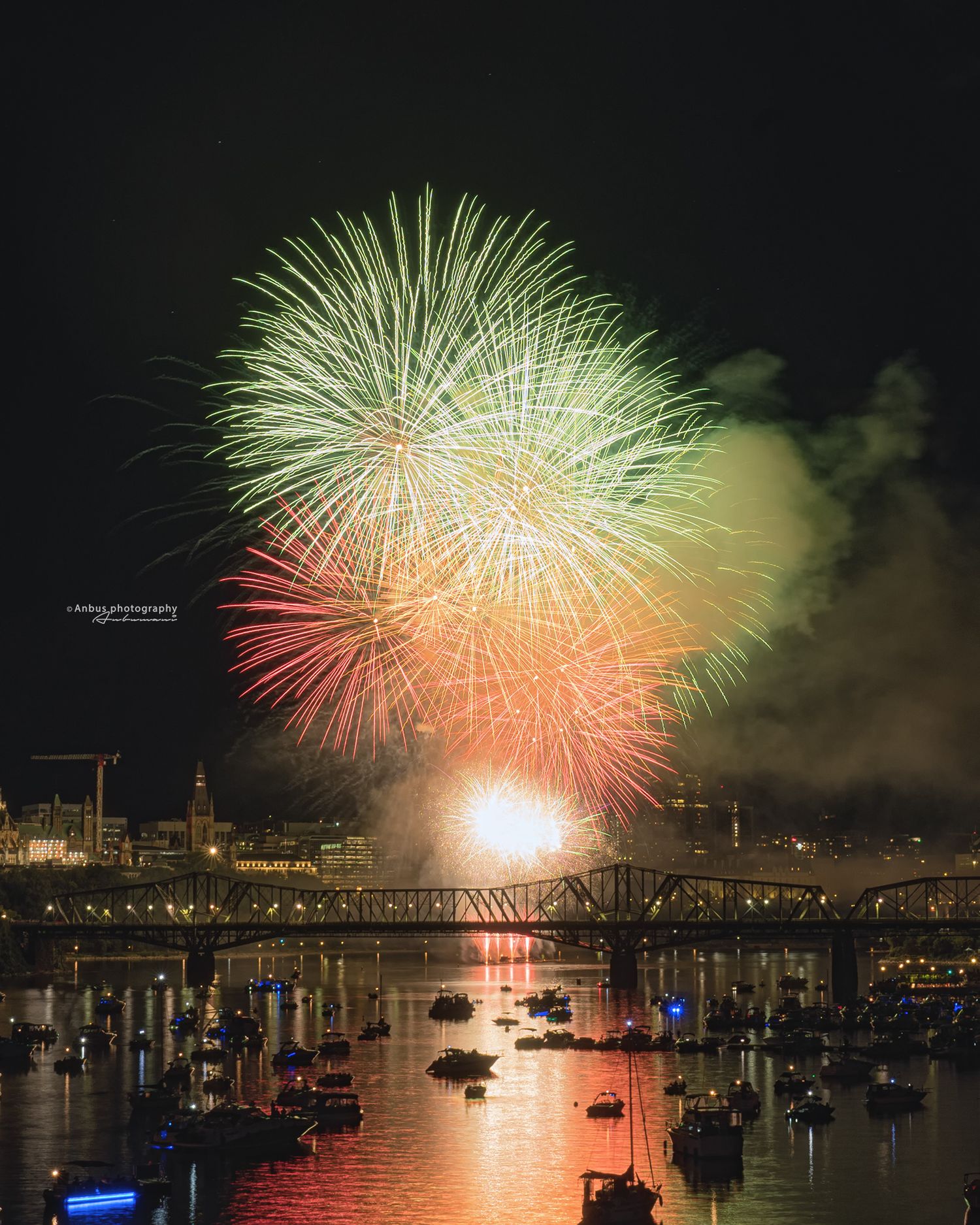 Colorful fireworks display illuminates the night sky over a harbor with boats and a bridge silhouetted against the water.