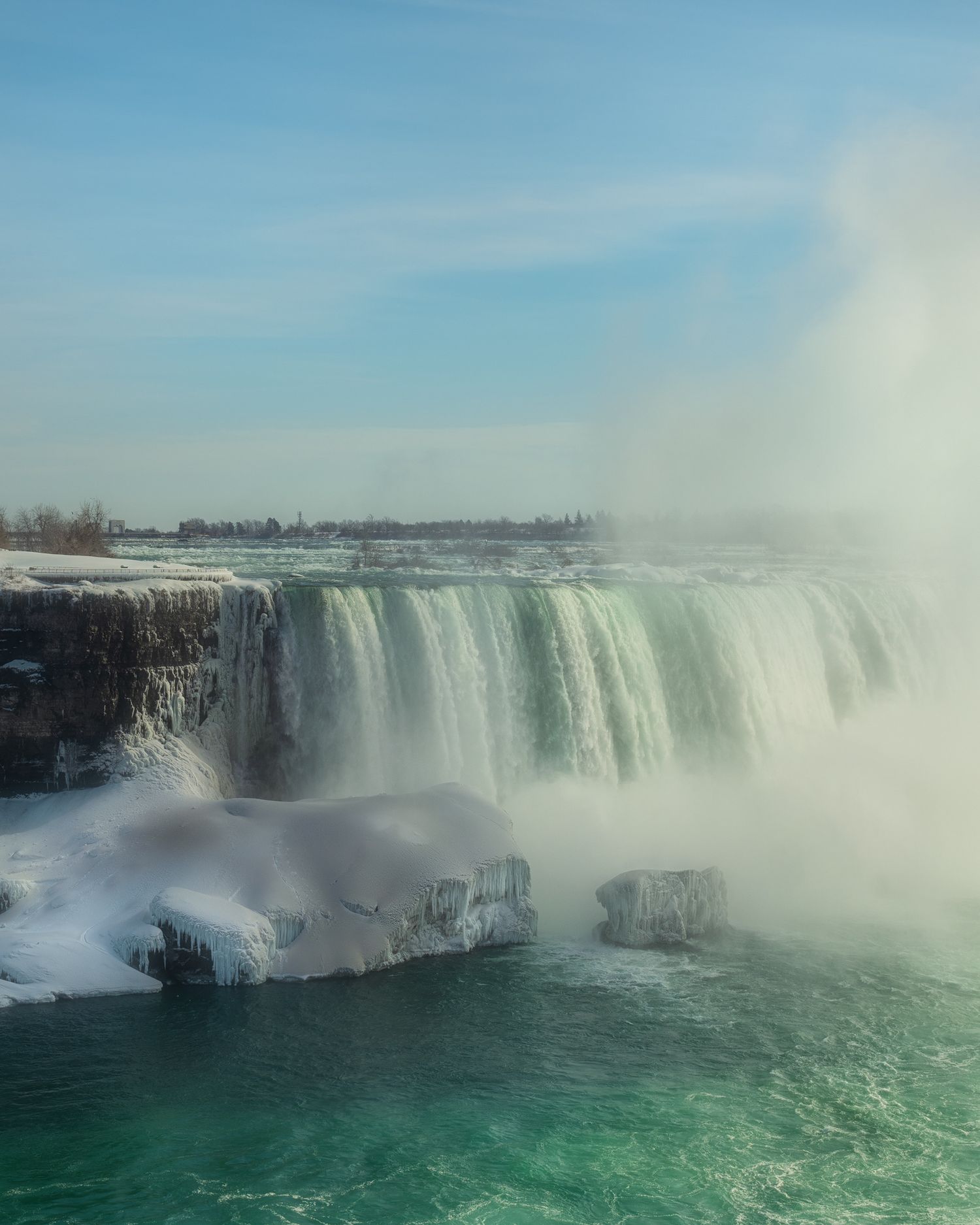 Majestic winter view of Niagara Falls with cascading water and turquoise pool below against a pale blue sky.