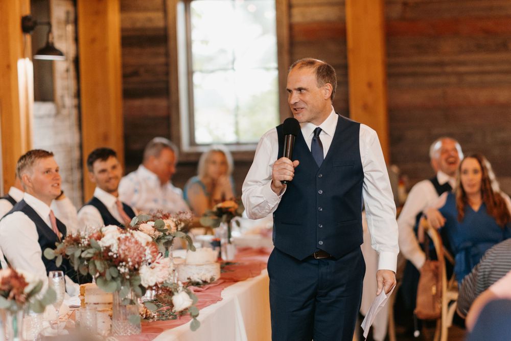 Groomsman in navy vest delivers wedding toast at floral-decorated head table in rustic barn reception.