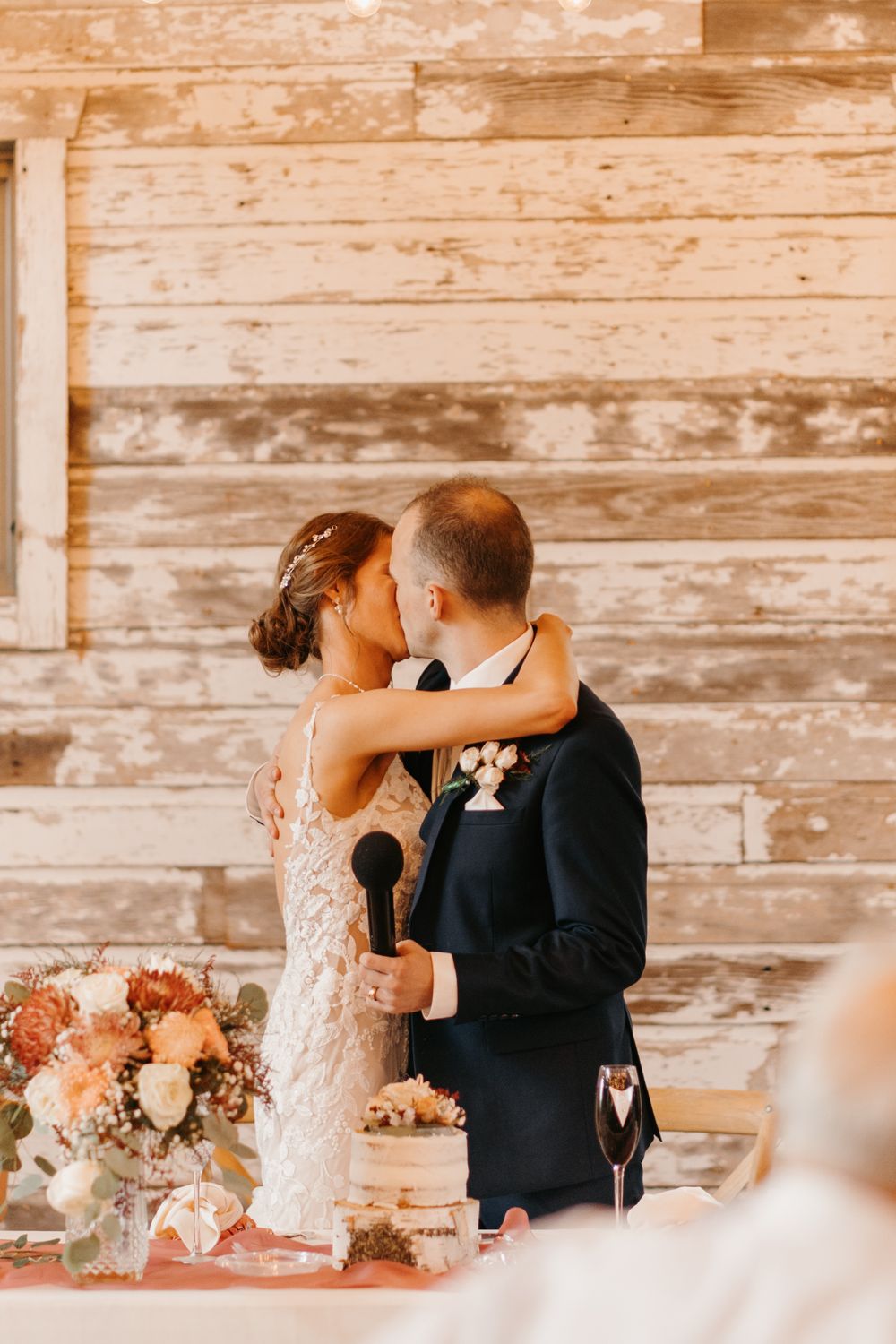 Bride and groom sharing a kiss at rustic barn wedding sweetheart table with floral arrangements.
