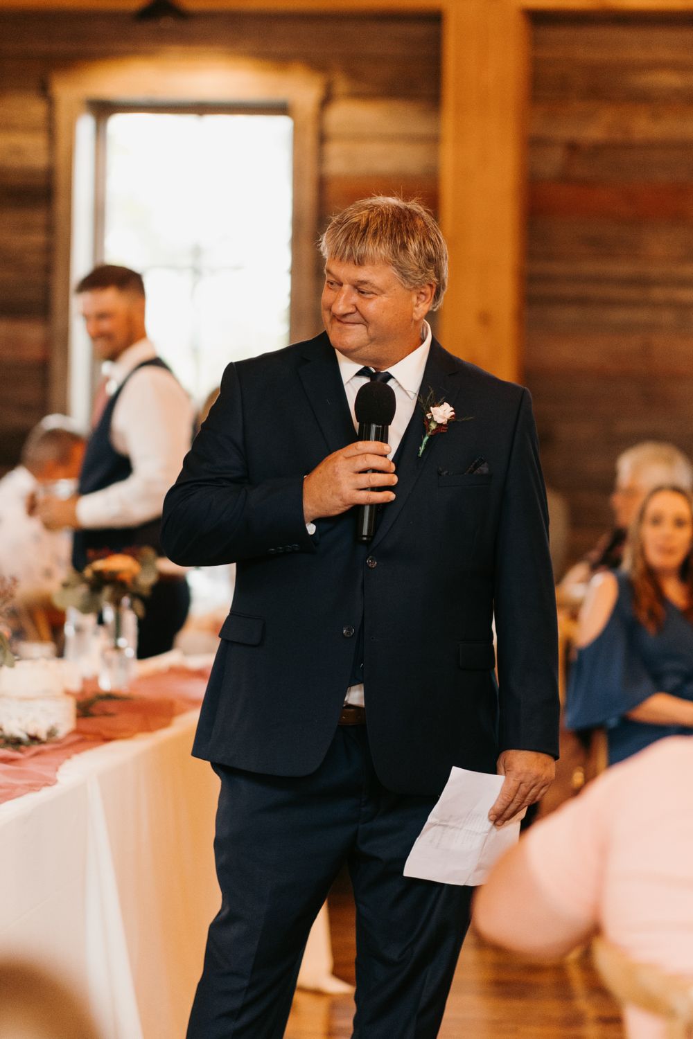 Father of bride smiles while giving wedding toast holding microphone and notes in rustic barn venue.
