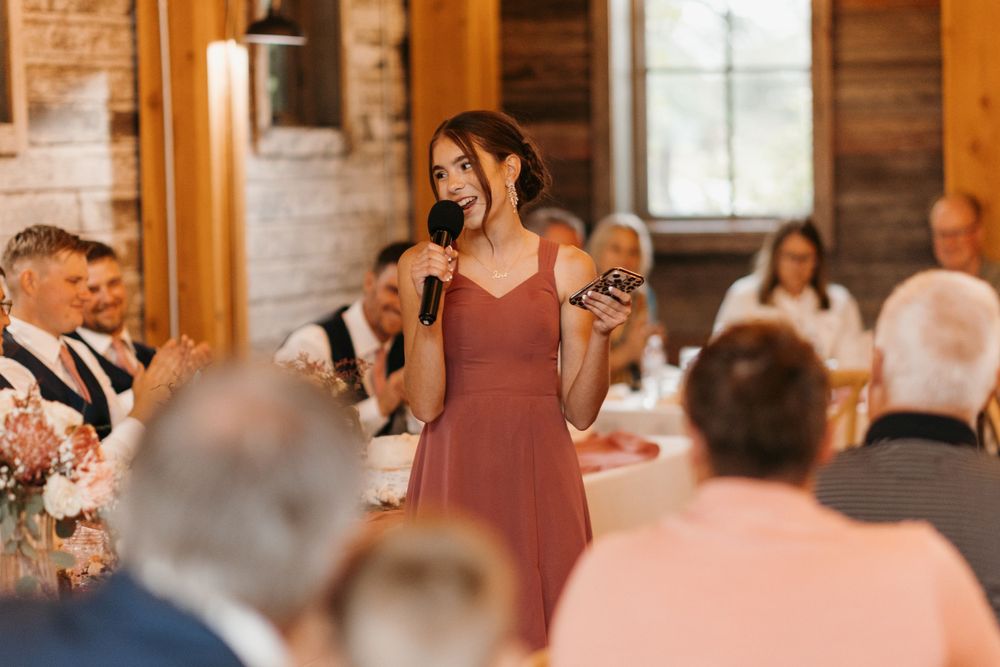 Bridesmaid in mauve dress delivering a wedding toast with microphone to seated guests in a rustic barn venue.