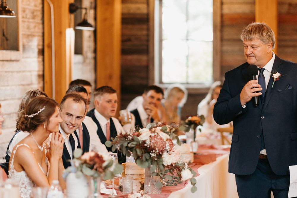 Father delivers touching wedding toast to laughing bride and groom at floral-adorned reception table.