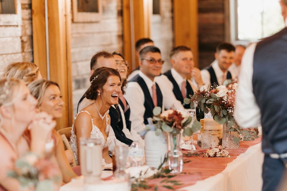 Bride laughs joyfully during wedding speeches at elegantly decorated head table in rustic barn venue.