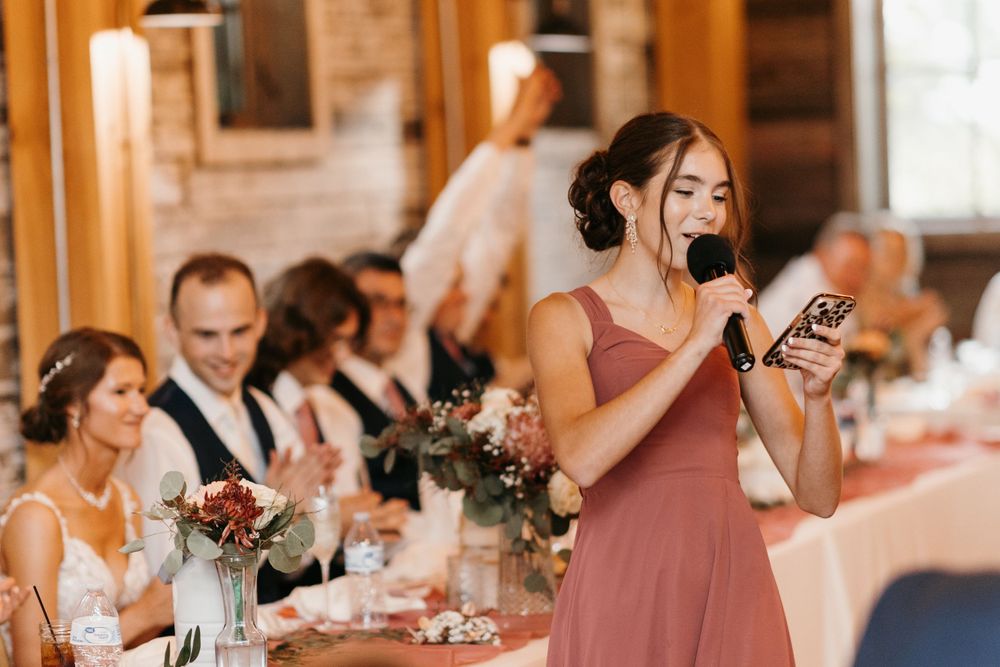Bridesmaid in dusty rose dress giving a wedding toast while holding a microphone and phone at reception.