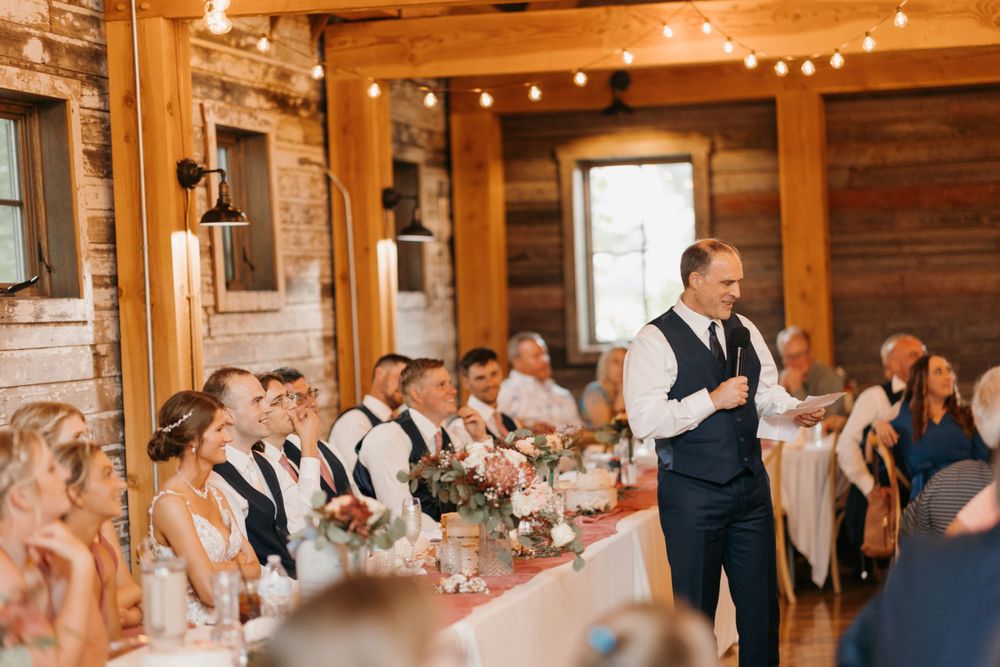 Man in navy suit gives wedding speech to bridal party at long decorated table in barn venue.