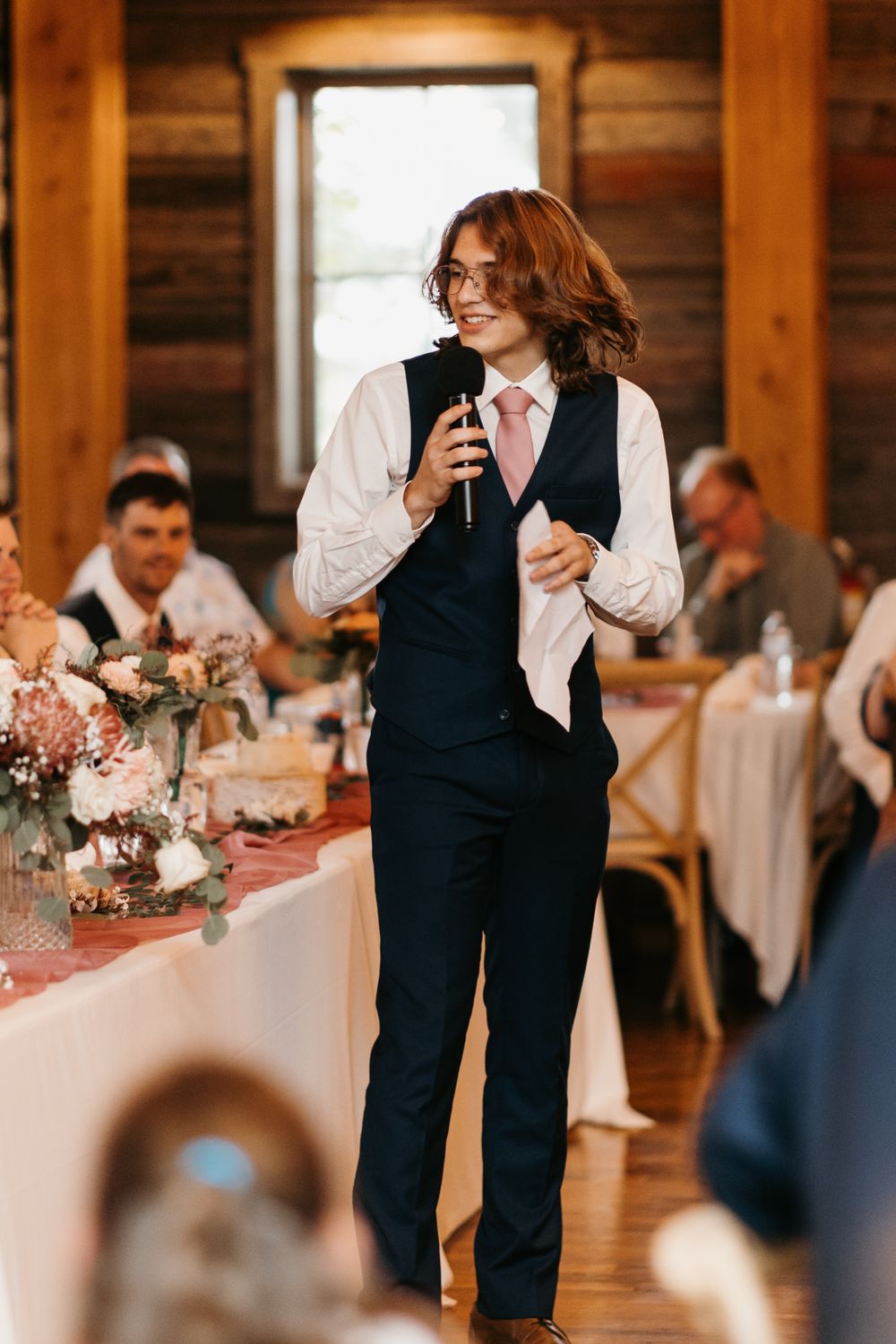 Young man in black vest and pink tie giving a wedding toast while holding a microphone at a rustic barn reception.