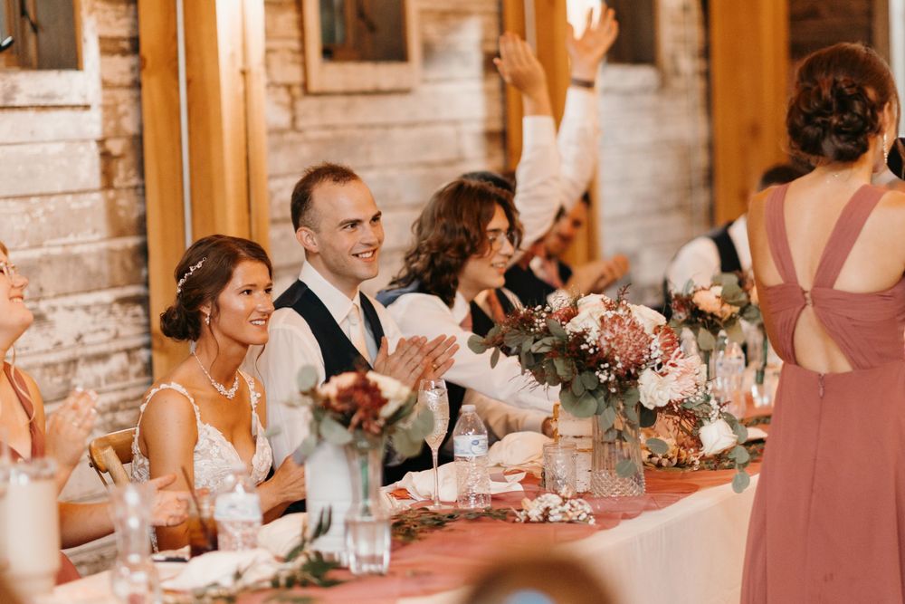 Bride and groom smiling and clapping at head table as bridesmaid raises hand during wedding reception toast.