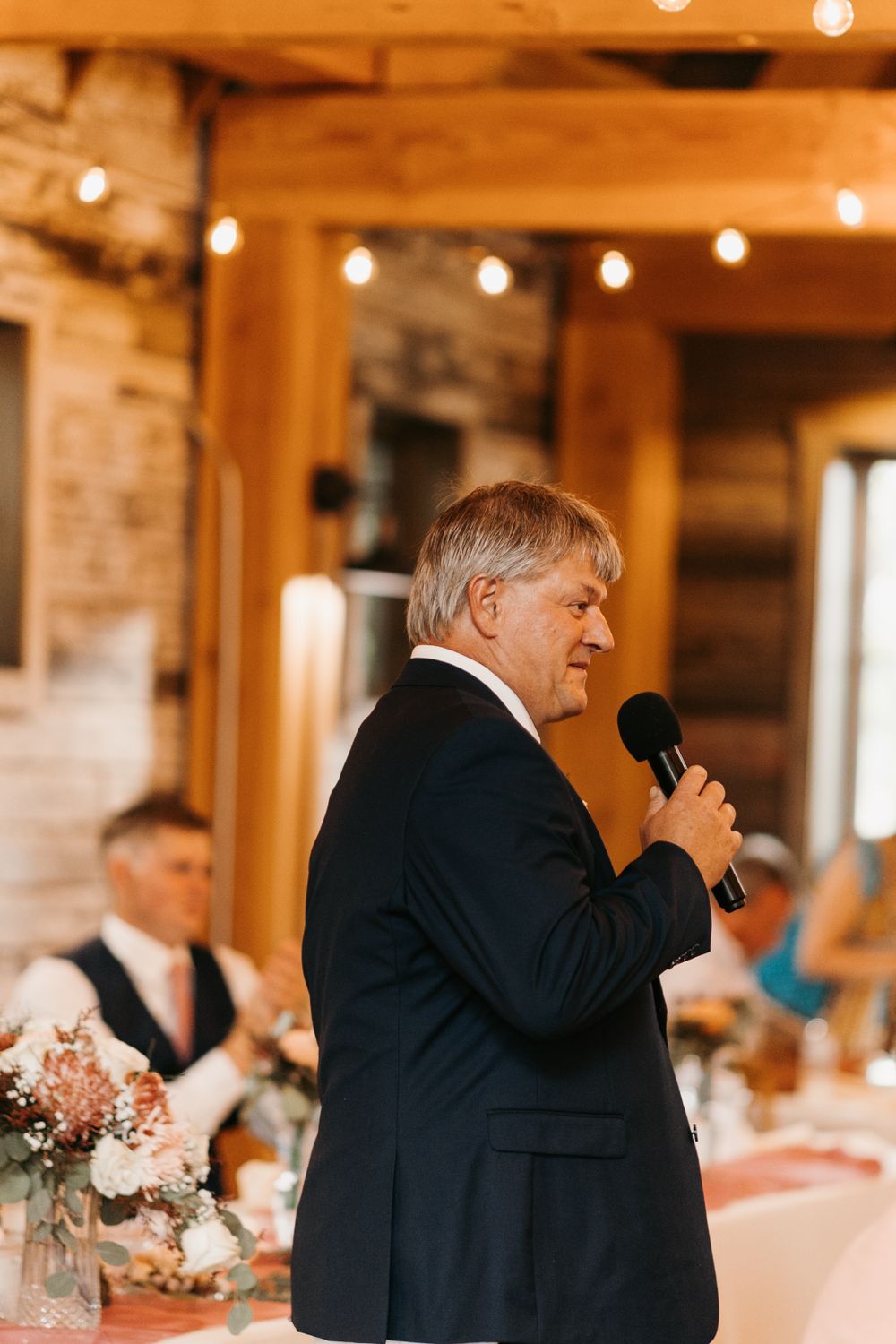 Older man in dark suit delivers heartfelt wedding speech into microphone at elegant barn reception.