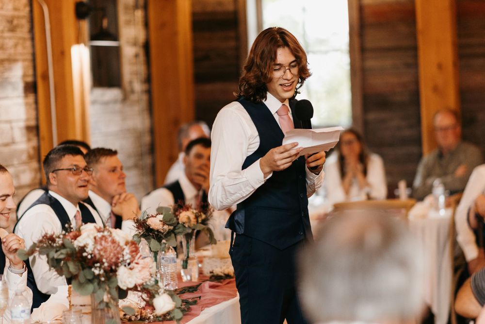 Person in black vest reading a heartfelt wedding speech to smiling guests at a rustic barn venue reception.