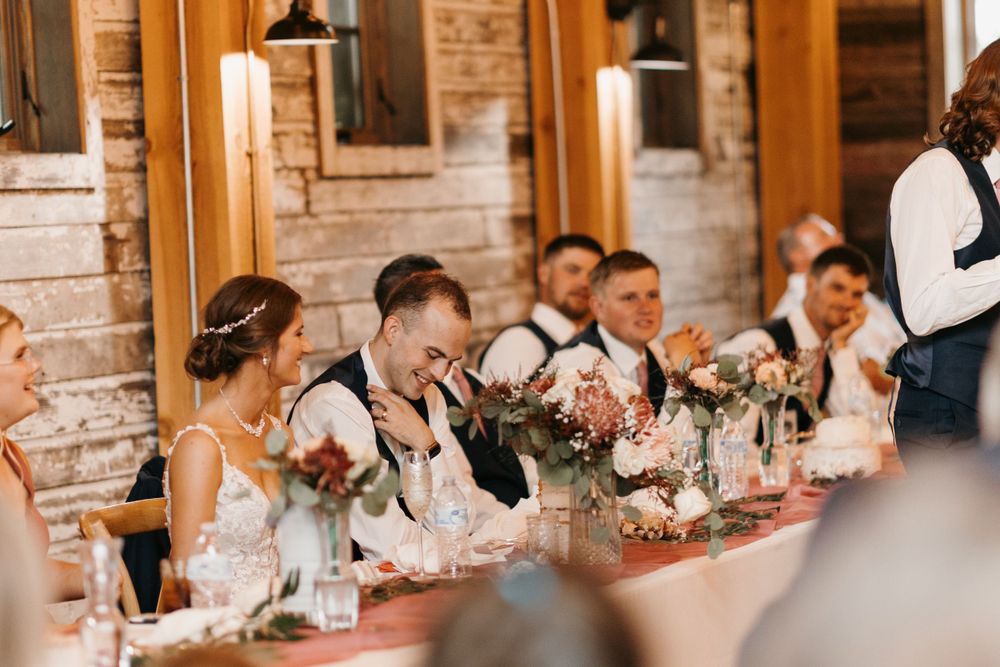Wedding party laughing and reacting joyfully at the head table during reception toasts in a rustic barn.
