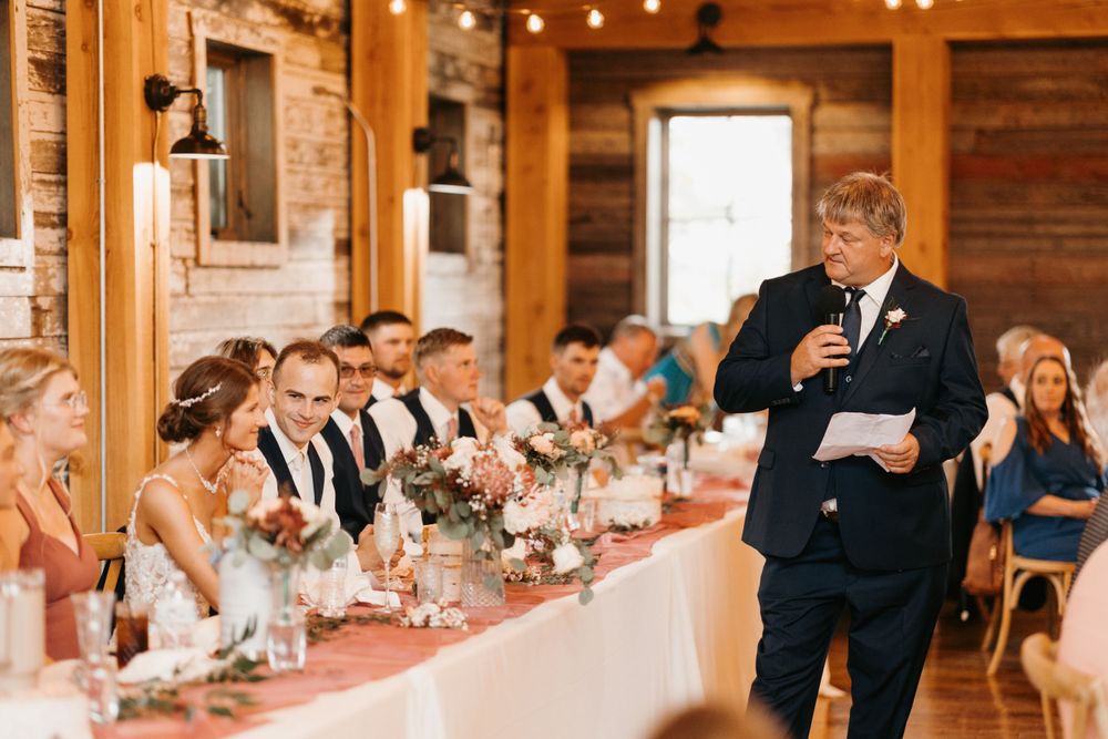 Man in navy suit reads wedding speech to bridal party seated at decorated head table in barn.