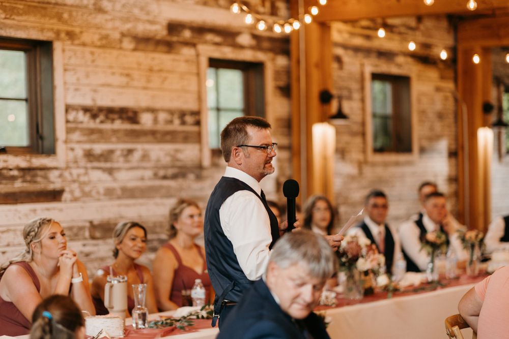 Best man in navy vest gives emotional wedding speech to bridal party at rustic barn reception.