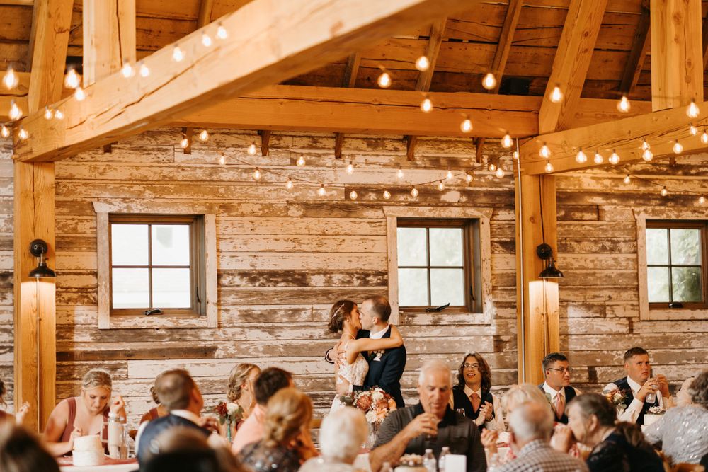 Couple shares first dance in rustic barn wedding venue adorned with string lights and wooden walls.