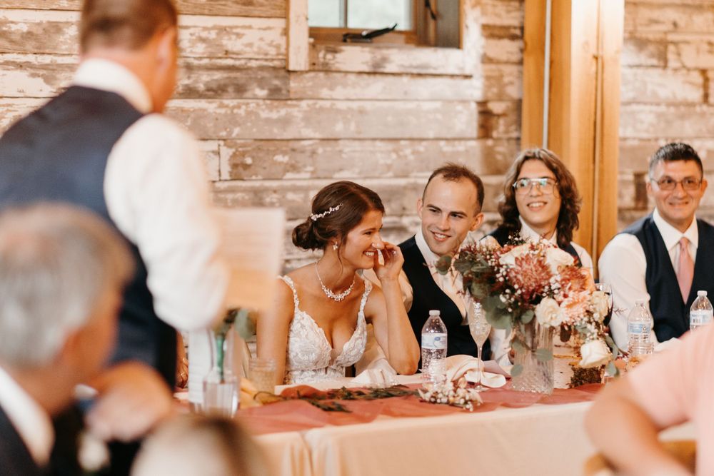Bride wipes tears while groom smiles during heartfelt wedding toast at rustic barn reception.