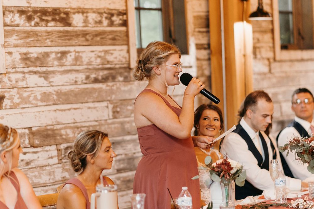Bridesmaid in dusty rose dress laughing while giving a wedding speech into a microphone at barn reception.