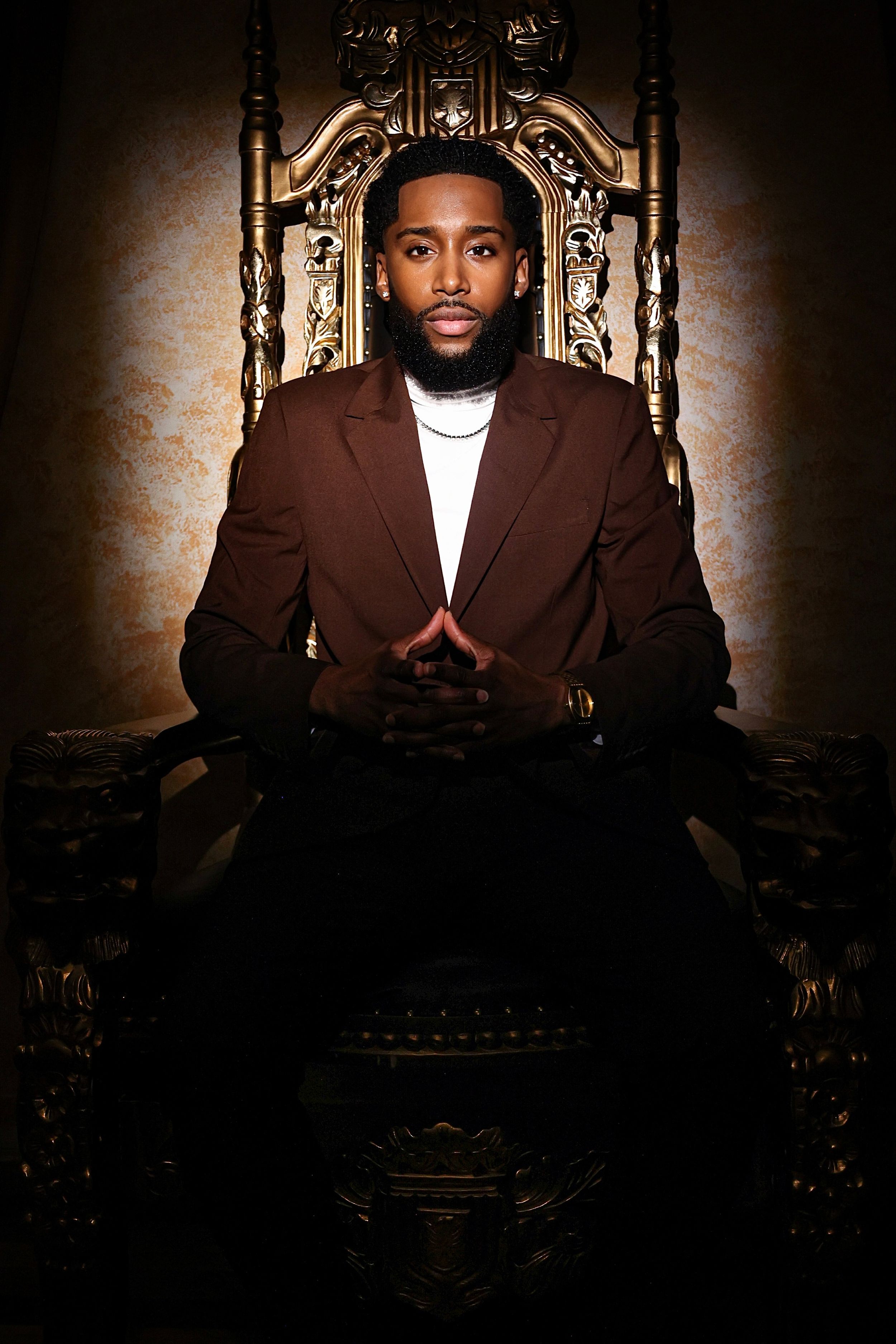 Dramatic portrait of person in brown suit seated on ornate throne chair.