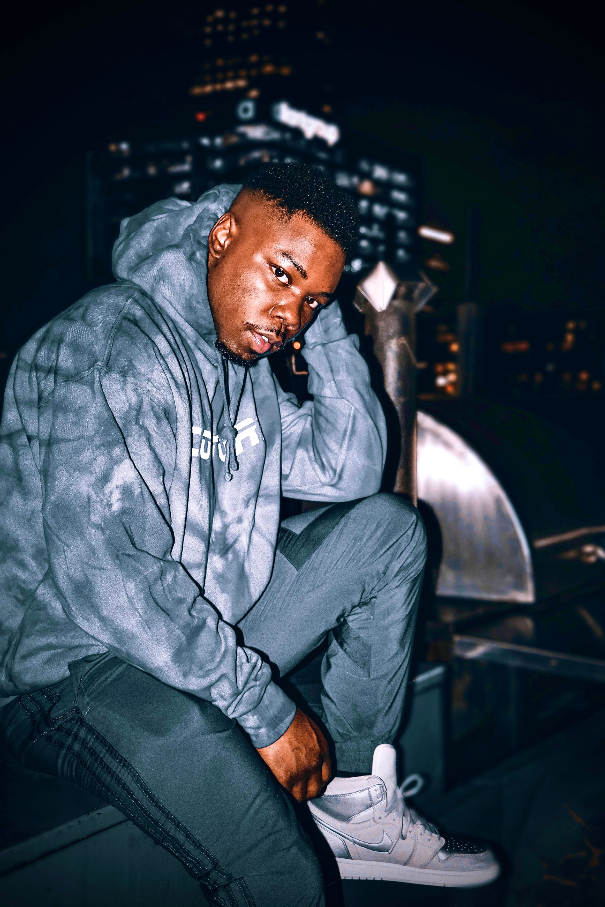 Nighttime urban portrait of person in grey hoodie and sneakers sitting thoughtfully against city backdrop.