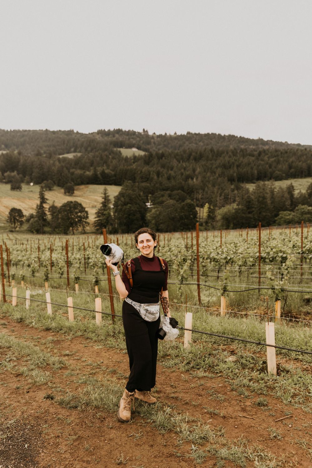 Person holding camera equipment while standing in vineyard with rolling hills in background.