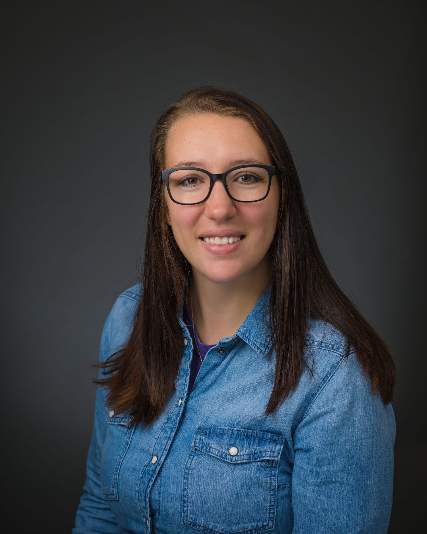 Professional headshot of person in denim shirt and glasses against dark background.