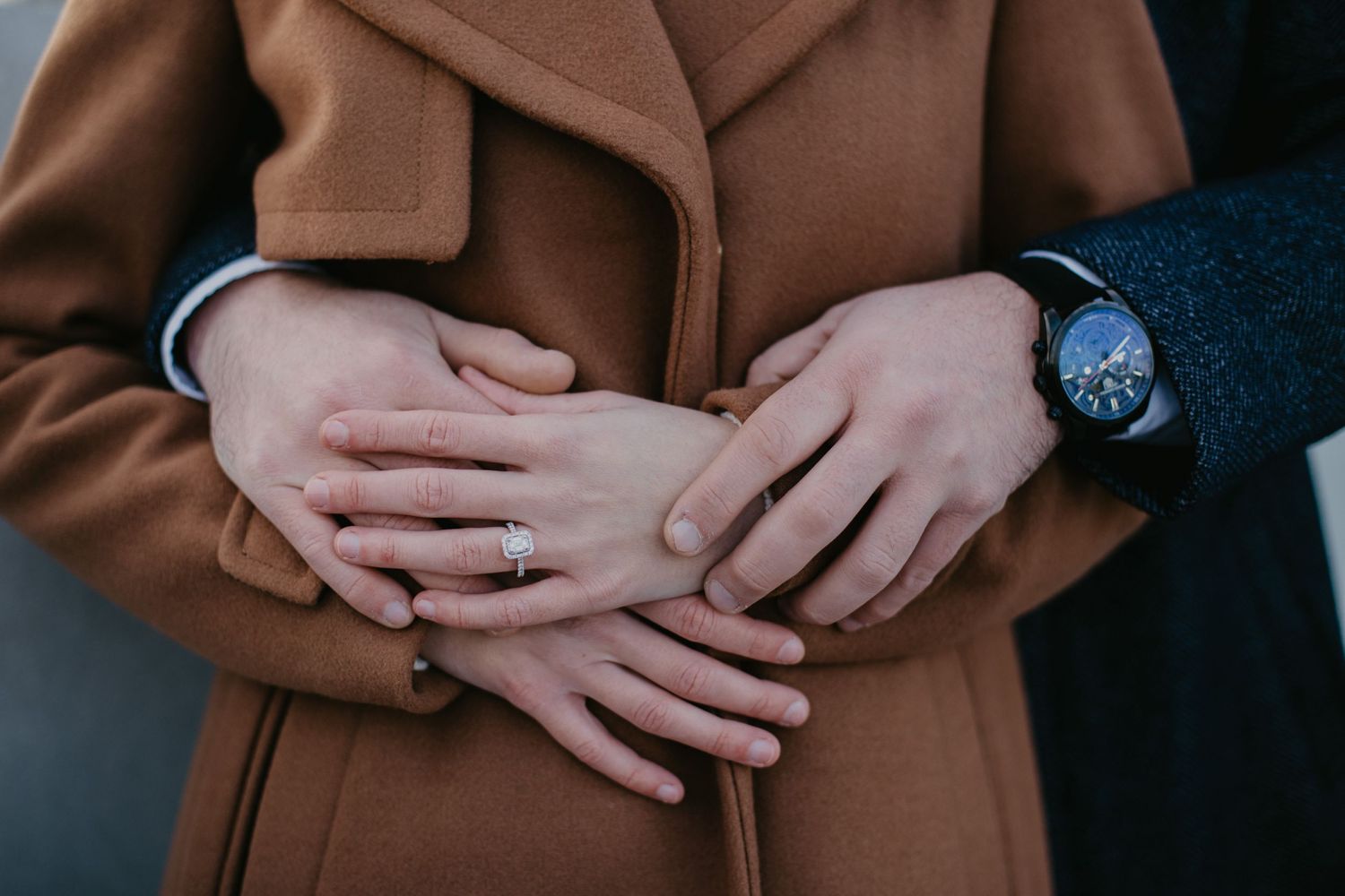 Close up of hands showing engagement ring with person wearing brown coat and watch.