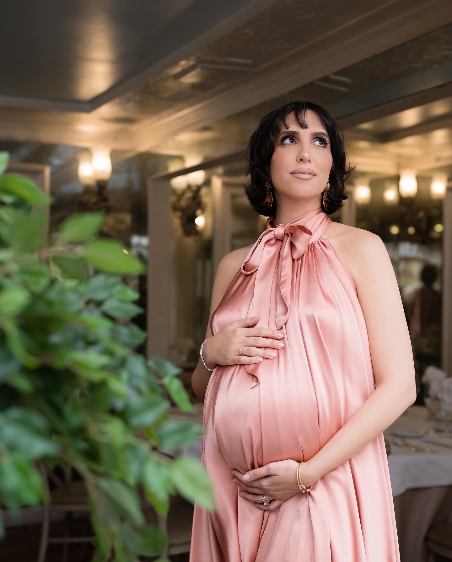 An expectant mother in a flowing peach-colored halter dress poses elegantly against an indoor backdrop with greenery.