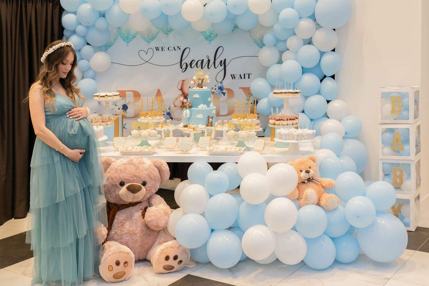 Blue and white balloon arch decorates a baby shower dessert table with teddy bears and treats.