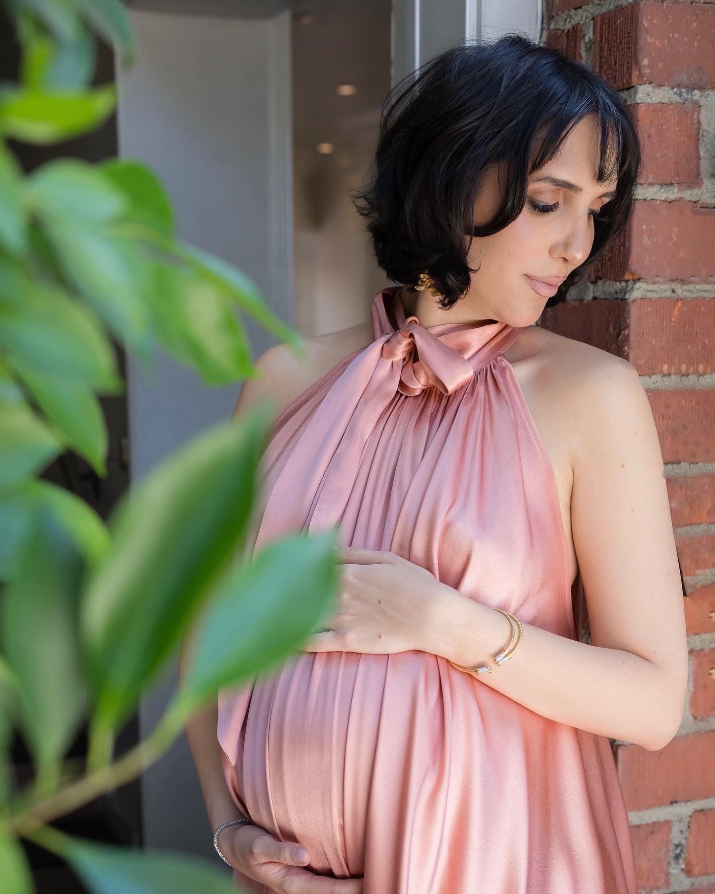 Side profile of person in pink dress standing against brick wall with greenery.